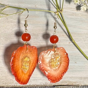 May include: A pair of handmade earrings featuring a strawberry slice design. Each earring has a gold-colored hook, a red bead, and a resin-coated strawberry slice with gold flakes. The earrings are set against a light wooden background.