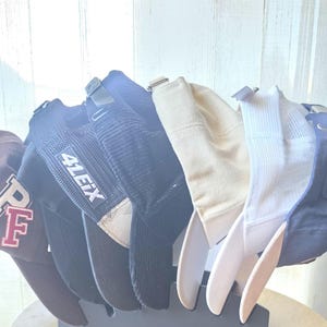 Könnte beinhalten: Eine Reihe von sechs Baseballkappen auf einem dunkelgrauen Kunststoffständer. Die Kappen umfassen eine kastanienbraune mit weißem Text, eine schwarze mit dem Text 41EIX, eine schwarze Cordkappe, eine beige Kappe, eine weiße Netz-Truckerkappe und eine marineblaue Kappe.