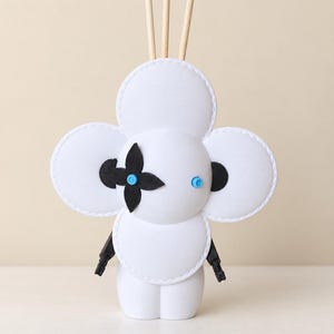 Puede incluir: Una figura decorativa blanca con forma de trébol y bordes cosidos, que presenta un acento floral negro de cuatro pétalos a la izquierda, un pequeño detalle circular azul a la derecha, brazos negros articulados y tres palos de madera finos.