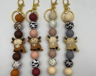 May include: Four keychains with gold-colored clasps and rings. Each keychain features a brown Highland cow charm, paired with a series of colorful beads in shades of brown, peach, and black and white patterns. The keychains are designed for attaching to keys or bags.