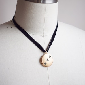 May include: A gold locket necklace with a black velvet choker. The locket has a round shape and features a design of three small butterflies with black accents.