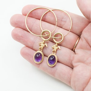 May include: Gold hoop earrings with oval, purple gemstones. Each earring features a small gold cherub figure above the gemstone. The earrings are held in a hand, showcasing their design and color.