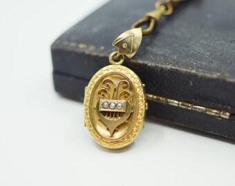 Op de afbeelding: Een goudkleurig ovaal medaillon met een sierlijk ontwerp, met een hart en drie kleine parels. Het medaillon hangt aan een delicate ketting. Het medaillon ligt op een donker, getextureerd oppervlak.