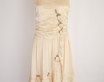 Op de afbeelding: Een vintage crèmekleurige jurk met een V-hals en mouwloos ontwerp. De jurk heeft een gerimpeld lijfje, een zwierige rok en bloemaccenten. De jurk wordt op een mannequin gepresenteerd.