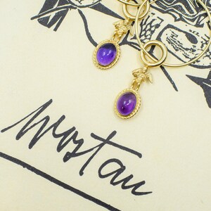May include: Gold hoop earrings with oval, purple gemstone pendants. Each pendant features a small gold figure above the gemstone. The earrings rest on a cream-colored surface with black illustrations and handwritten text.