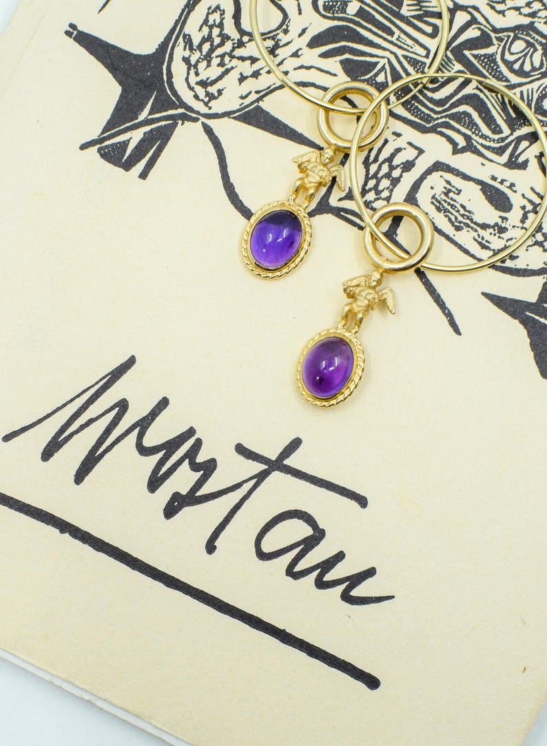 May include: Gold hoop earrings with oval, purple gemstone pendants. Each pendant features a small gold figure above the gemstone. The earrings rest on a cream-colored surface with black illustrations and handwritten text.