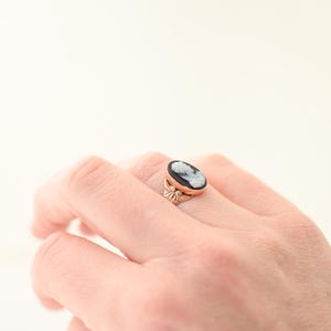 Op de afbeelding: Een goudkleurige ring met een ovale camee met een wit profiel tegen een zwarte achtergrond. De ringband heeft een decoratief bloemmotief. De ring zit aan een hand.