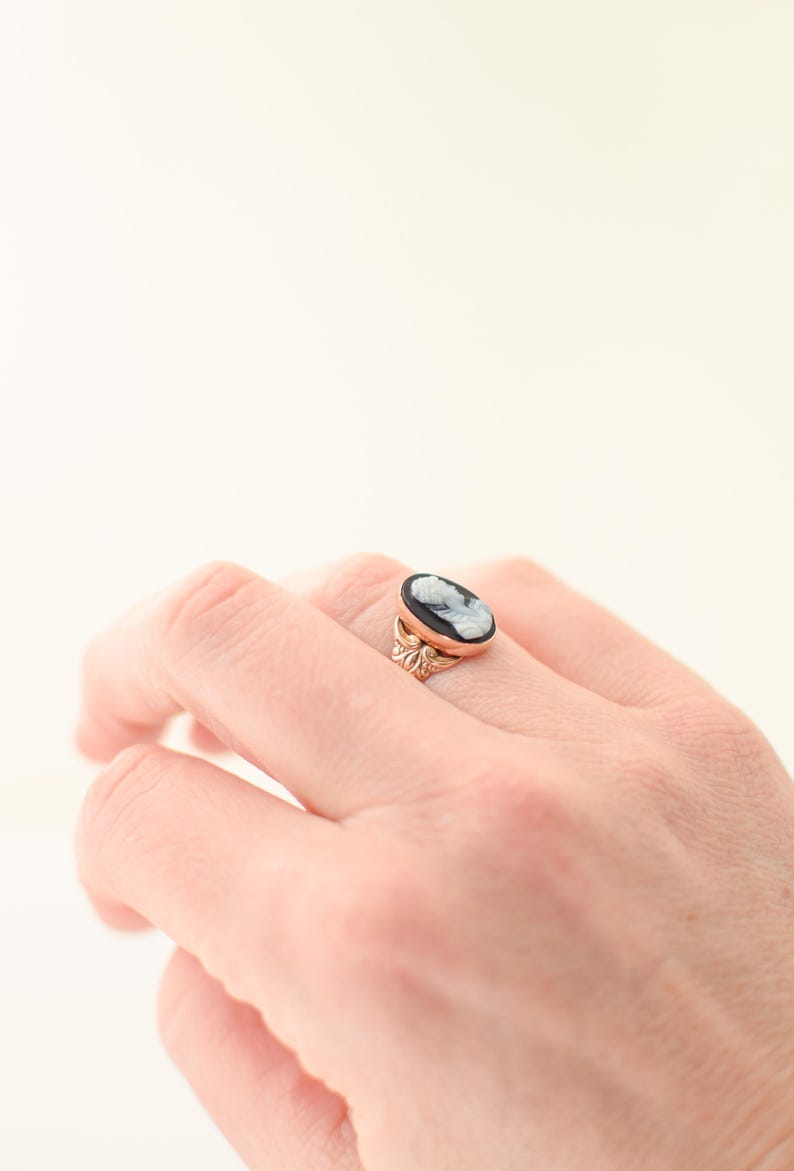 Op de afbeelding: Een goudkleurige ring met een ovale camee met een wit profiel tegen een zwarte achtergrond. De ringband heeft een decoratief bloemmotief. De ring zit aan een hand.