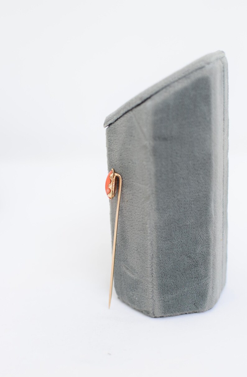 May include: A gold pin with a red coral stone set in a gold frame. The pin is on a gray velvet display stand.