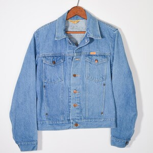 May include: Light blue denim jacket with a classic collar, button closure, and two chest pockets. The jacket has a vintage look, with a small orange tag on the chest pocket. The jacket is hanging on a wooden hanger.