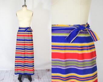 Op de afbeelding: Een lange, gestreepte rok in rood, blauw, geel en groen. De rok heeft een wikkelstijl met een blauwe strik in de taille. De stof lijkt getextureerd en de rok wordt op een mannequin getoond. De lengte is ongeveer 100 cm.