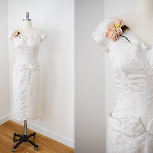May include: A white, short-sleeved dress with a floral pattern and a sweetheart neckline. The dress has a fitted bodice and a skirt with pockets. A pink flower is pinned to the dress.