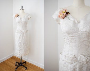 May include: A white, short-sleeved dress with a floral pattern and a sweetheart neckline. The dress has a fitted bodice and a skirt with pockets. A pink flower is pinned to the dress.