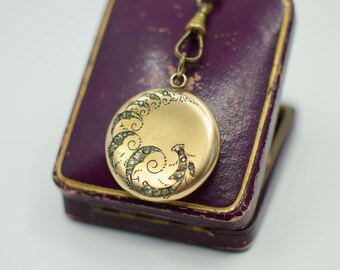 May include: A gold-colored, round locket with an ornate, swirling design, accented with small, sparkling stones. The locket hangs from a chain and rests on a purple, rectangular box with a gold trim. The box is slightly open.