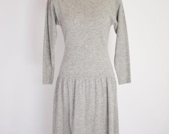 May include: A light gray, long-sleeved dress with a round neckline and a fitted bodice. The dress features a gathered skirt that falls to the mid-calf. The dress is displayed on a mannequin against a white background.