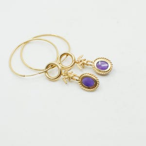 May include: Gold-toned hoop earrings with a dangling oval-shaped purple stone. The earrings feature a small figure above the stone and a twisted rope design around the stone's edge. The hoops are thin and circular.