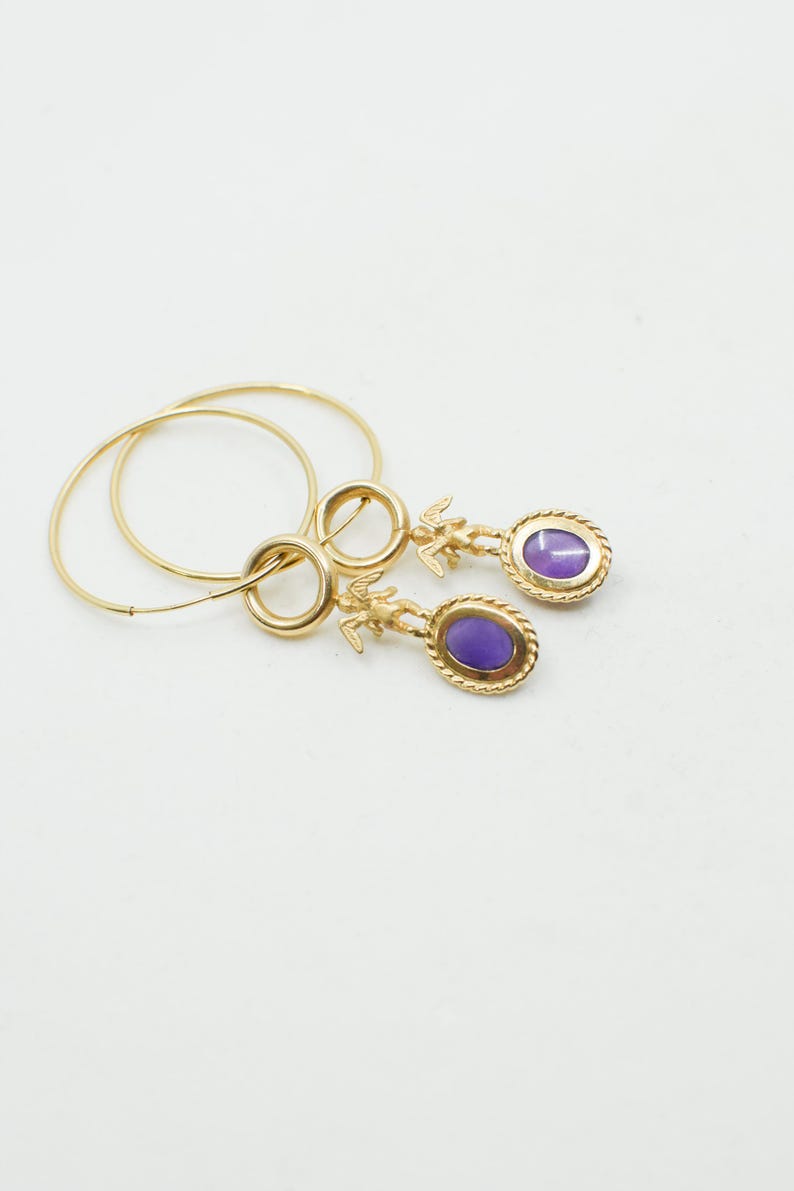 May include: Gold-toned hoop earrings with a dangling oval-shaped purple stone. The earrings feature a small figure above the stone and a twisted rope design around the stone's edge. The hoops are thin and circular.