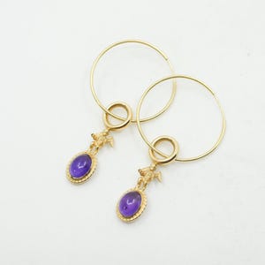 May include: A pair of gold-colored hoop earrings. Each earring features a small, oval, purple gemstone pendant with a gold-colored cherub figure above it. The earrings are displayed against a white background.