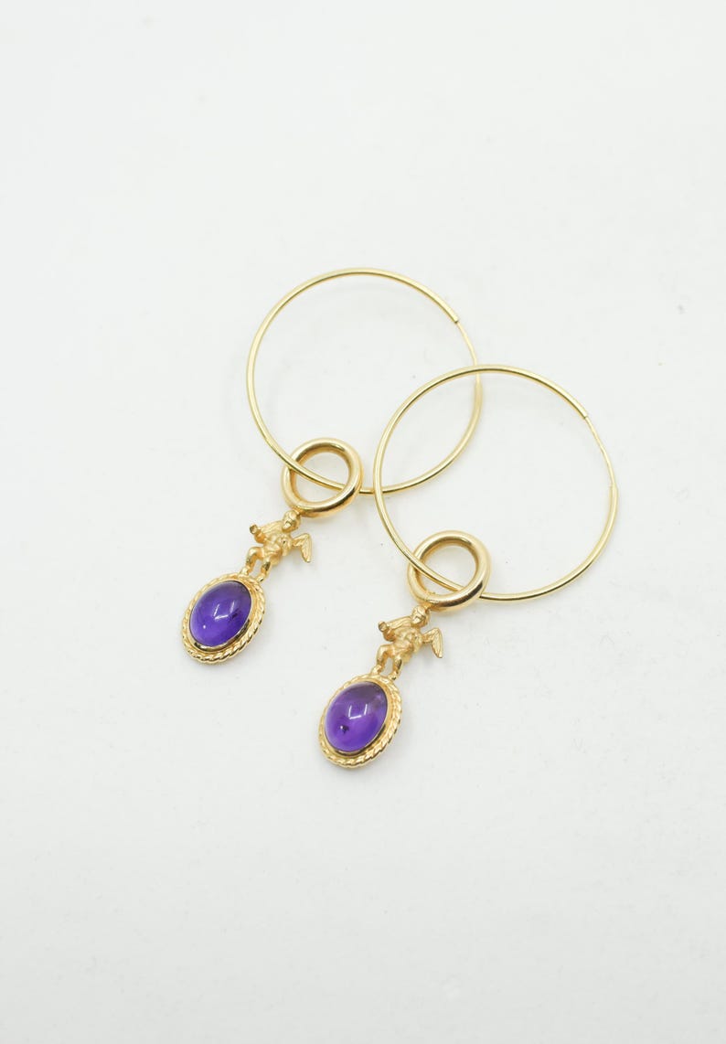 May include: A pair of gold-colored hoop earrings. Each earring features a small, oval, purple gemstone pendant with a gold-colored cherub figure above it. The earrings are displayed against a white background.