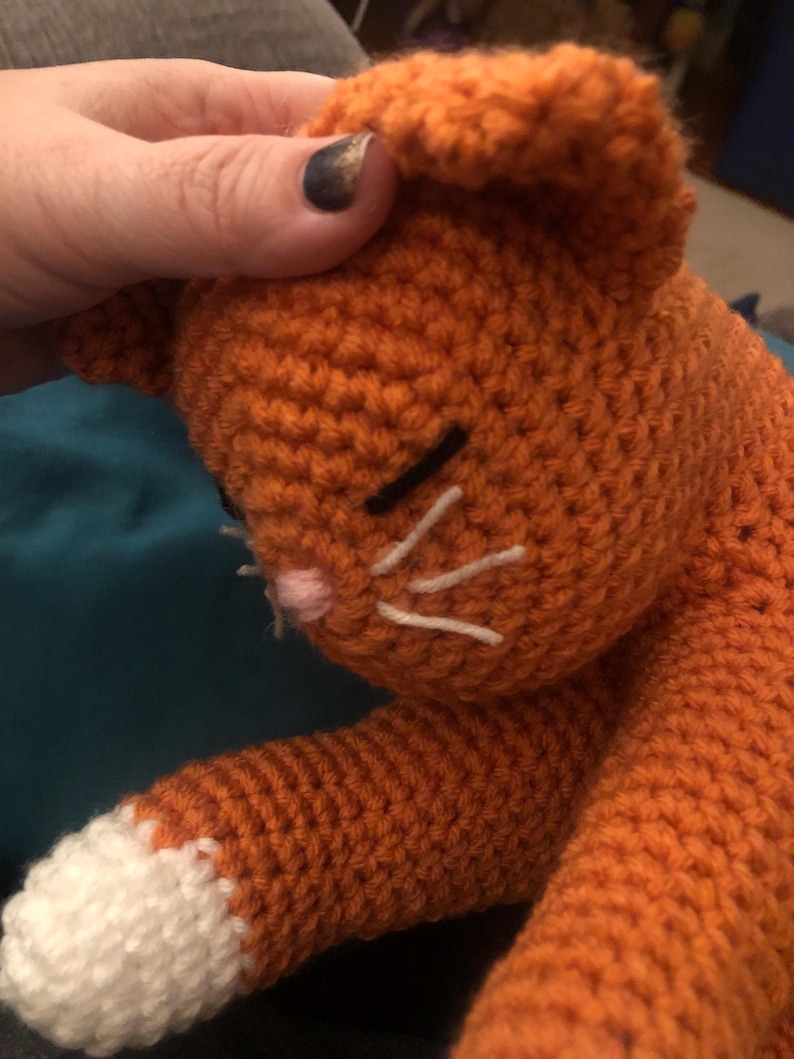 May include: A crocheted orange cat with white paws and a pink nose. The cat has black eyes and white whiskers.
