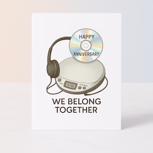 Puede incluir: Una tarjeta de felicitación con una ilustración de dibujos animados de un reproductor de CD portátil y auriculares. Un CD con "Happy Anniversary" está encima. El texto "We Belong Together" está debajo del reproductor.
