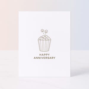 Puede incluir: Una tarjeta de felicitación blanca con un dibujo lineal marrón de una magdalena con palomitas de maíz y las palabras "HAPPY ANNIVERSARY" debajo. El diseño es simple y minimalista.