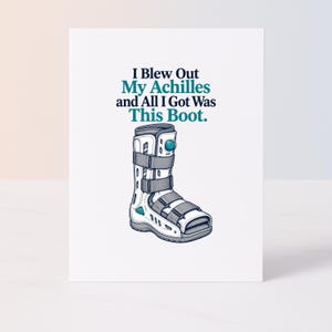May include: A white greeting card featuring a graphic illustration of a medical walking boot with grey straps and a teal air valve. Above the boot, the text reads: I Blew Out My Achilles and All I Got Was This Boot. The design is centred on the card.