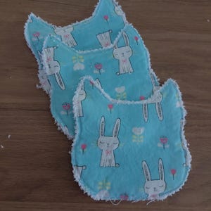 Puede incluir: Tres baberos azules para bebé con un estampado de conejitos y flores. Los baberos tienen una forma única y ribete blanco. El diseño del conejito incluye un lazo y está rodeado de flores rosas y blancas.