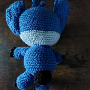 Puede incluir: Un peluche azul tejido a mano con grandes orejas, una franja azul claro y detalles negros. El juguete est&aacute; sobre una superficie de madera. El peluche es un personaje de una popular pel&iacute;cula de animaci&oacute;n.