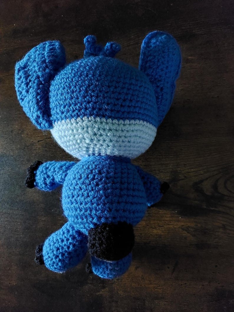Puede incluir: Un peluche azul tejido a mano con grandes orejas, una franja azul claro y detalles negros. El juguete est&aacute; sobre una superficie de madera. El peluche es un personaje de una popular pel&iacute;cula de animaci&oacute;n.