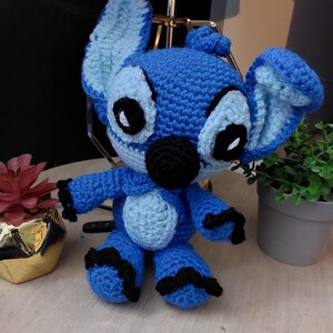 Puede incluir: Mu&ntilde;eco Stitch azul hecho a mano con ganchillo, con orejas grandes, ojos negros y nariz negra. El mu&ntilde;eco tiene una barriga azul claro y detalles negros en brazos y pies. Est&aacute; sobre una superficie de madera.