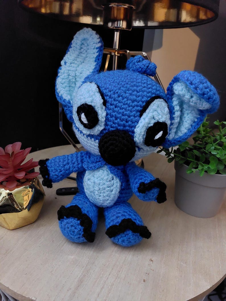 Puede incluir: Mu&ntilde;eco Stitch azul hecho a mano con ganchillo, con orejas grandes, ojos negros y nariz negra. El mu&ntilde;eco tiene una barriga azul claro y detalles negros en brazos y pies. Est&aacute; sobre una superficie de madera.
