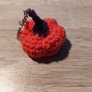 Könnte beinhalten: Ein handgefertigter Kürbis-Schlüsselanhänger aus Häkelarbeit. Der Kürbis ist orange mit einem dunkelvioletten Stiel. Der Schlüsselanhänger ist an einem silbernen Metallring und einer Kette befestigt. Der Kürbis hat einen Durchmesser von etwa 5 cm.