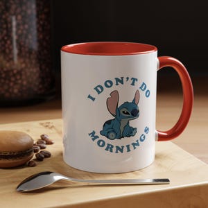 Puede incluir: Taza de cerámica blanca con interior y asa rojos. La taza presenta un personaje de dibujos animados azul y el texto "I DON'T DO MORNINGS" en azul. Una cuchara y un pastel están en primer plano.
