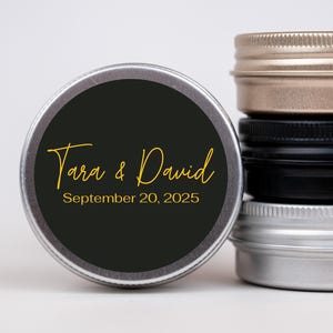 May include: A stack of four small, round metal tins with screw-on lids. One tin has a black label with gold text that reads "Tara & David September 20, 2025." The lids are silver, black, and gold.