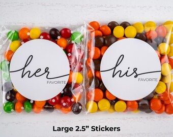 Peut inclure: Deux sacs transparents remplis de bonbons colorés. Chaque sac a un autocollant circulaire blanc avec les mots "her favorite" et "his favorite" en écriture noire. Le texte "Large 2.5" Stickers" est en bas.