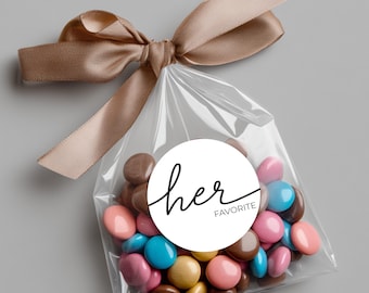 Peut inclure: Un sac transparent rempli de bonbons colorés, noué avec un ruban de satin beige. Un autocollant circulaire blanc sur le sac indique "her favorite" en écriture noire.