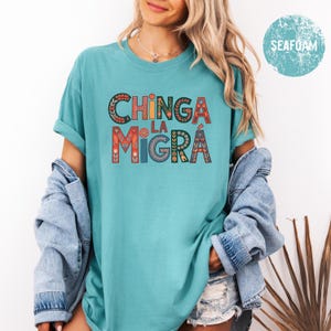 May include: Seafoam green t-shirt with the text "CHINGA LA MIGRA" in colorful, decorative lettering. The shirt has rolled-up sleeves and is paired with a denim jacket. The design features floral and geometric patterns.
