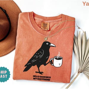 May include: A yam-colored Comfort Colors crew neck t-shirt featuring a black raven holding a coffee mug. The text "HELLO DARKNESS MY OLD FRIEND" is printed below the raven. A gold necklace and a brown hat are also visible.