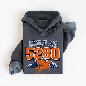 May include: Dark gray hooded sweatshirt with the text "BUILT AT 5280" in white and orange. Below the text is a graphic of a winged horse and mountains. The sweatshirt is made of a soft material.