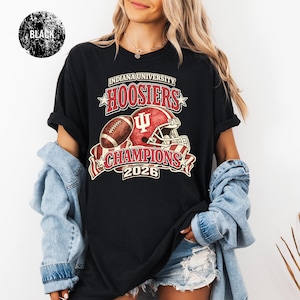 May include: Black t-shirt featuring a graphic design for Indiana University Hoosiers. The design includes a football, a red helmet, and the text "HOOSIERS CHAMPIONS 2026". The shirt is worn by a person with long blonde hair.
