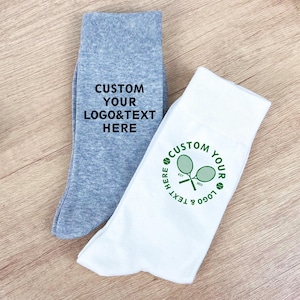 Könnte beinhalten: Zwei Paar personalisierte Socken, eine graue und eine weiße, werden auf einer Holzoberfläche gezeigt. Die graue Socke trägt den Text "CUSTOM YOUR LOGO & TEXT HERE". Die weiße Socke zeigt ein Tennis-Logo mit dem Text "CUSTOM YOUR LOGO & TEXT HERE".