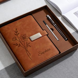 Pode incluir: Um diário de couro castanho com um design floral e o nome "Sophia" gravado na capa. O diário está numa caixa de presente com uma caneta e uma caneta stylus. O diário tem um fecho prateado.
