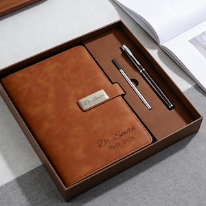 Puede incluir: Un diario de cuero marrón con un cierre plateado y un juego de bolígrafo y bolígrafo a juego, presentado en una caja marrón. El diario está grabado con el nombre "Dr. Smith" y "Ph.D. 2026".