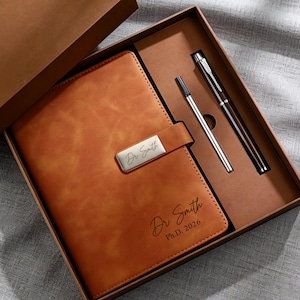 May include: A brown leather journal and two pens are presented in a brown gift box. The journal has a silver clasp with the name "Dr. Smith" engraved on it. The journal also has the name "Dr. Smith" and "Ph.D. 2026" engraved on the front.