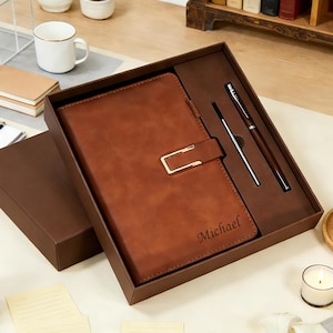 Könnte beinhalten: Ein braunes Lederjournal mit goldenem Verschluss, präsentiert in einer passenden Geschenkbox. Das Journal enthält einen Stift und einen Stylus. Der Name "Michael" ist auf dem Journal eingraviert. Das Set wird auf einer hellen Oberfläche präsentiert.