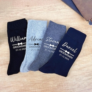 May include: Four pairs of groomsmen socks in black, light grey, medium grey, and navy blue. Each sock has a name, bow tie graphic, and date printed in white. The socks are arranged on a wooden surface.
