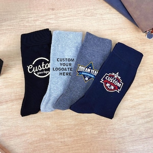 Op de afbeelding: Vier paar aangepaste sokken in zwart, lichtgrijs, donkergrijs en marineblauw. Elke sok heeft een uniek ontwerp, inclusief de tekst "Custom your logo" en "Dream Team". De sokken worden gepresenteerd op een houten oppervlak.