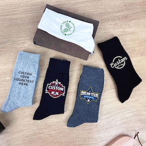 Puede incluir: Vista aérea de calcetines personalizados en varios colores, incluyendo gris, azul marino y negro, con logotipos y textos personalizados. Se ve una mascarilla blanca con un logotipo de tenis, junto con un pequeño frasco de perfume y una caja de regalo.