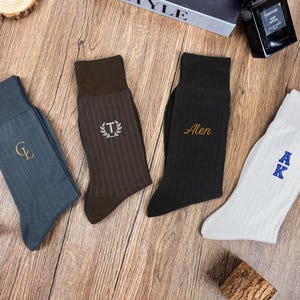 May include: Four pairs of ribbed dress socks in various colors: teal, brown, black, and white. Each sock features embroidered initials or a name in a contrasting color. The socks are arranged on a wooden surface.
